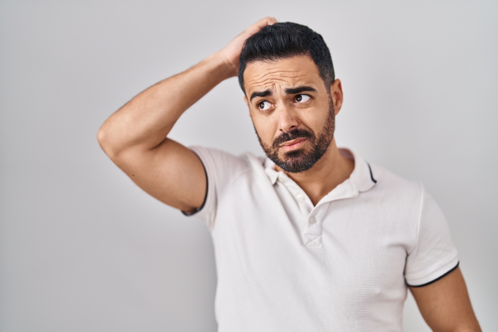 Young hispanic man with beard wearing casual clothes over white background confuse and wondering about question. uncertain with doubt, thinking with hand on head. pensive concept.